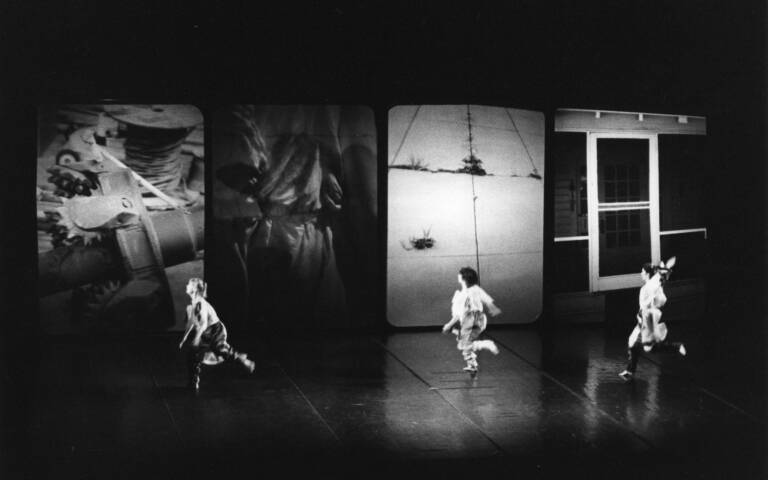 Three dancers in a dramatic stage setting with four large, abstract screens in the background displaying various images